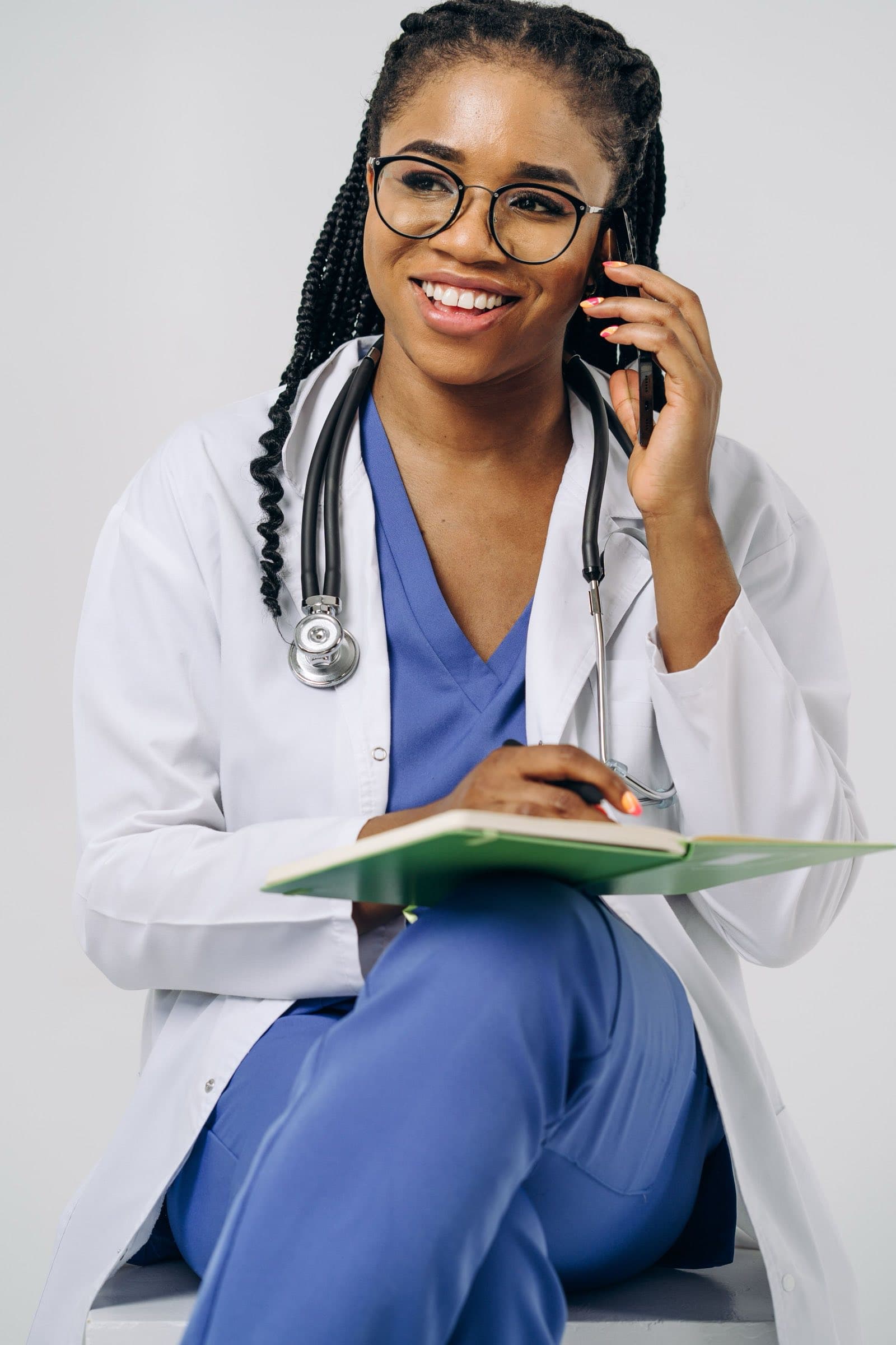 A doctor in a white coat and scrubs, smiling on a phone call with her notes